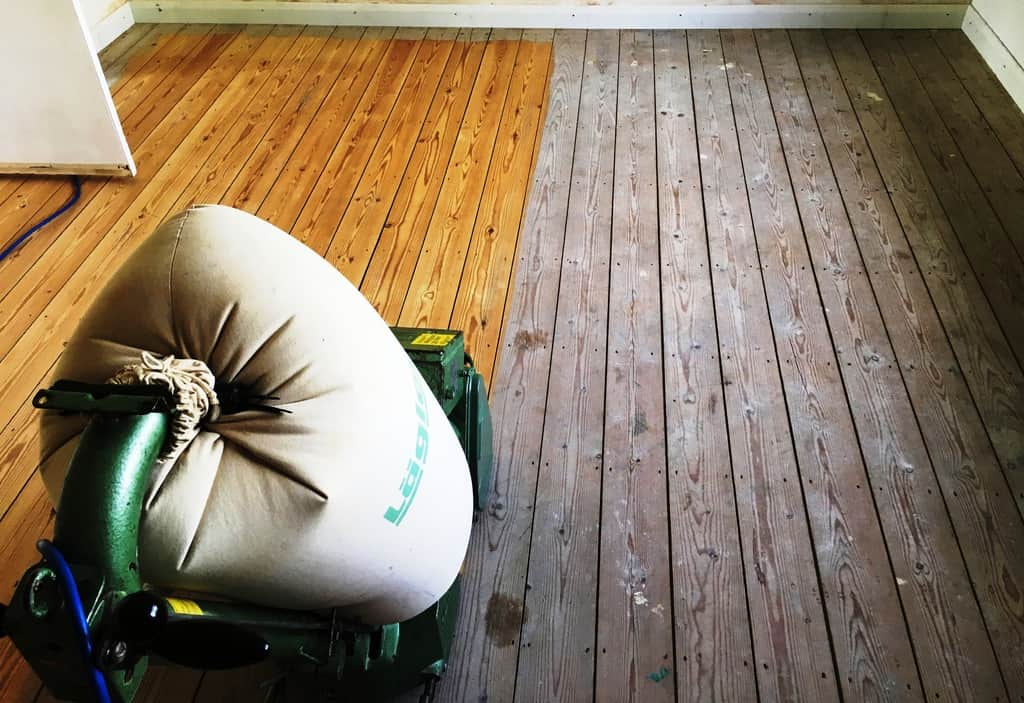 A half-sanded wooden floor, with a Lagler sanding machine in the foreground, showcasing the dramatic transformation from a worn gray surface to a beautifully restored golden wood finish.