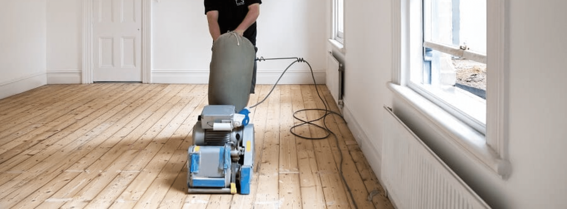 Floor sander in action, smoothing wooden surface for refinishing. Protective gear including goggles, mask, and ear muffs worn. The sander's powerful motor and rotating abrasive pad create a smooth, even finish. Fine dust particles visible in the air. Preparation and precision are key to achieving professional results.
