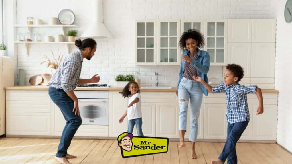 A joyful family dancing in their beautifully renovated kitchen with hardwood floors by Mr Sander® in Cudham, UK. The parents and two children hold hands and smile as they move around the bright, modern kitchen featuring white cabinets, open shelving, and a white brick backsplash. The high-quality wood flooring adds warmth and elegance to the space, highlighting the exceptional floor sanding and refinishing services provided by Mr Sander® in Bishopstone. The Mr Sander® logo is visible at the bottom of the image.