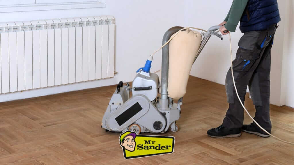 A man from Mr Sander® is sanding an oak wood floor in the living room of a home in Medstead, UK. using a Bona Belt (AMO110000.3) sanding machine. The Bona Belt features a 2.2 kW motor, 230 V voltage, 50 Hz frequency, 200 & 250 mm drum width, and a sanding belt size of 200 x 750 mm or 250 x 750 mm, with a total weight of 74 kg.