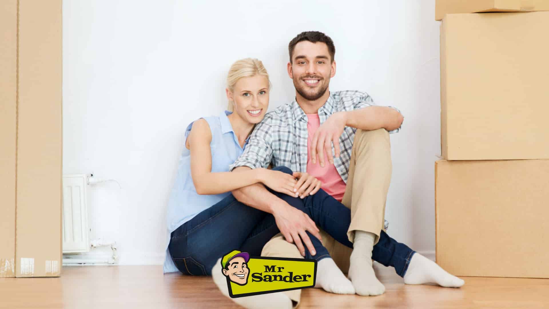 A happy couple sitting on the floor with moving boxes around them in Chadwell Heath, UK, with a Mr Sander® logo overlay. The image showcases a beautifully refinished wooden floor.