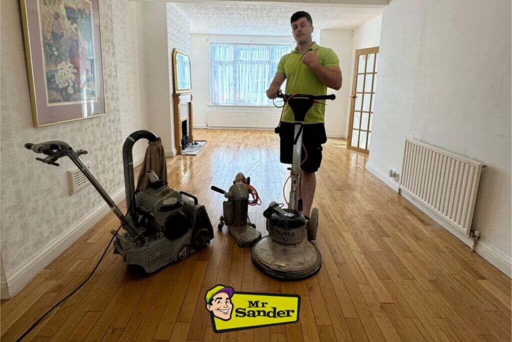 A professional floor technician from Mr. Sander stands with a sanding machine in a well-lit living room with hardwood flooring. Multiple sanding tools are lined up in front of him, showing the equipment used for floor sanding and refinishing. The room is bright, with a large window, light-colored walls, and minimal decor, highlighting the smooth and polished wooden floor. The Mr. Sander logo is displayed at the bottom center of the image.