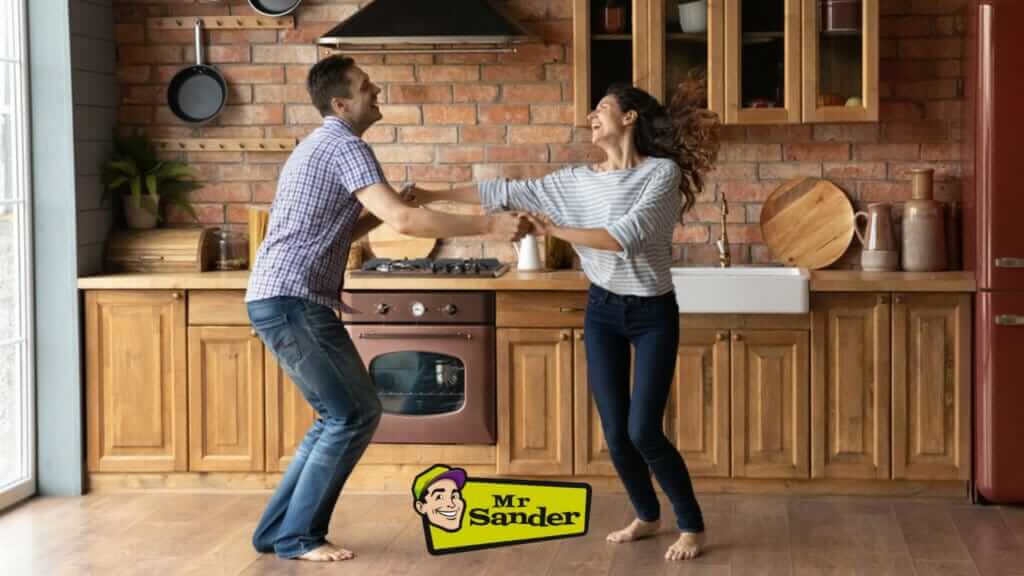 A joyful couple dances in their kitchen with a beautifully refinished oak plank floor in Catford, UK. The floor, expertly sanded and refinished by Mr Sander®, enhances the rustic charm of the kitchen with its brick wall, wooden cabinets, and modern appliances, creating a warm and inviting atmosphere.