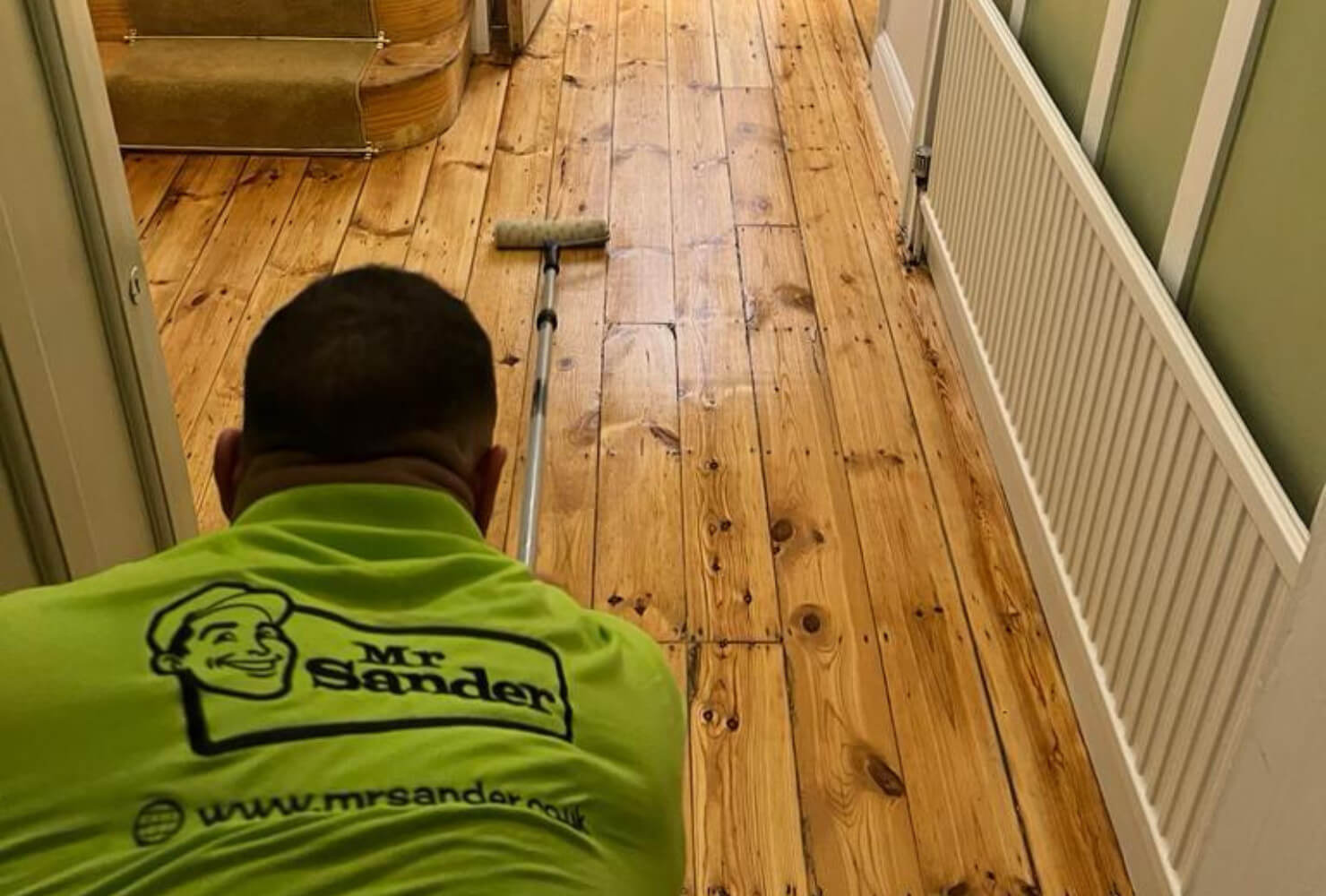 A Person from Mr. Sander, wearing a bright green uniform with the company logo, is seen from behind as he works on restoring a pine wood floor. He uses a roller to apply a finish, showcasing his expertise in floor restoration. The hallway leads to a staircase, with the freshly sanded and varnished floor extending throughout.