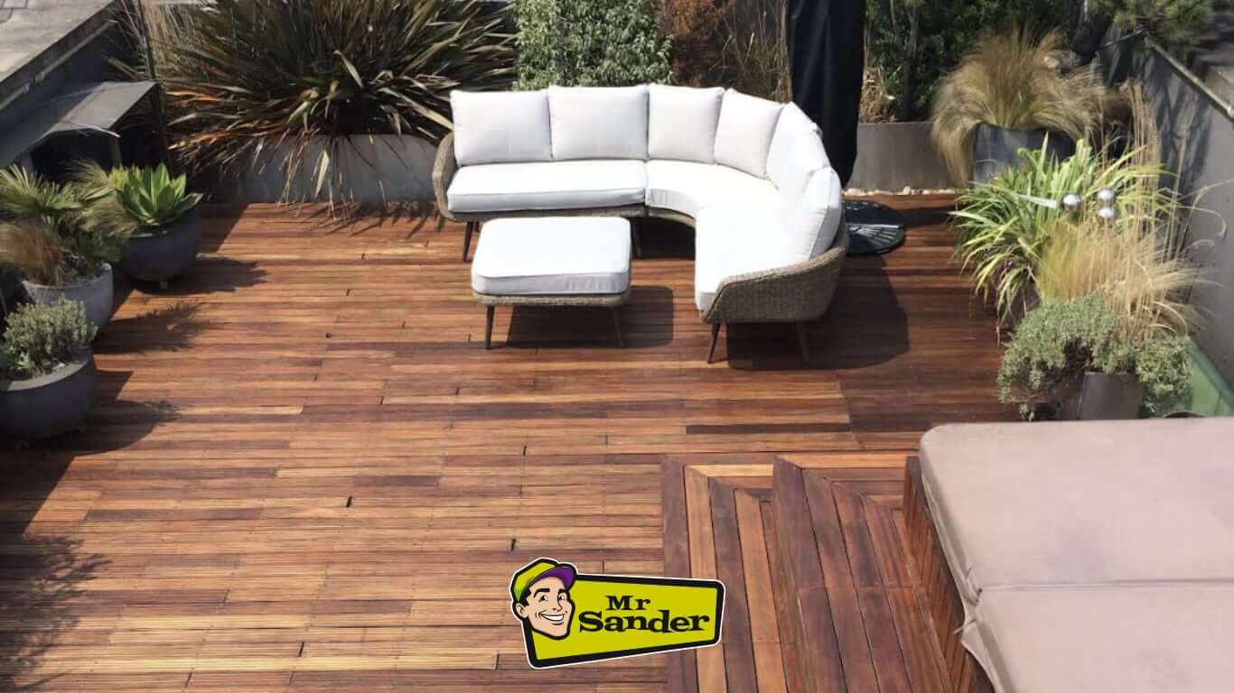 A spacious outdoor deck with a wooden floor, overlooking a cityscape. The deck features a curved sofa, potted plants, and a small table. The Mr. Sander logo is visible in the lower right corner.