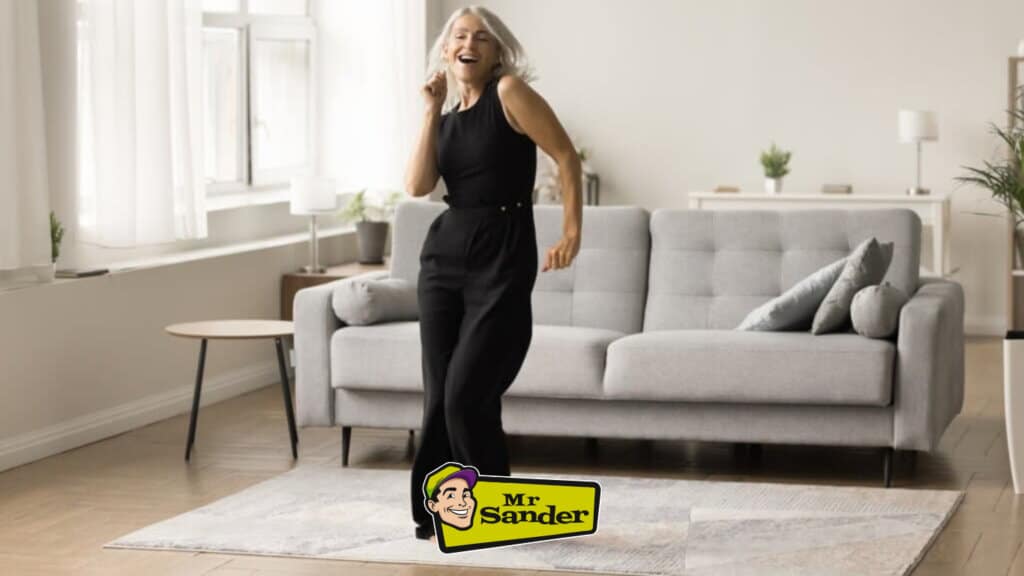 A joyful older woman in a black outfit dances in a stylish living room with a modern gray sofa. The room is illuminated by natural light streaming through large windows, highlighting the smooth and beautifully refinished wooden floors. The elegant parquet flooring adds warmth and character to the minimalistic decor. Floor sanding and refinishing done by West Thurrock