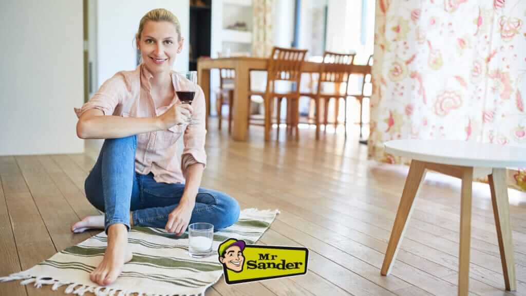 A woman sitting on a rug on a smoothly refinished light oak hardwood floor by Mr Sander in Bulphan, UK, enjoying a glass of wine.