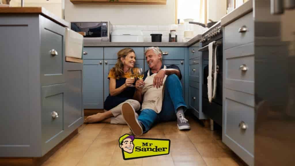 A happy couple sitting on the beautifully sanded and refinished kitchen floor, enjoying drinks. The modern kitchen in Lower Edmonton, UK, showcases the high-quality floor sanding and refinishing services provided by Mr Sander®.