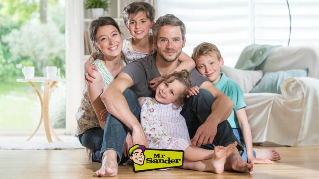 A happy family of five sitting together on a beautifully refinished wooden floor. The image showcases the quality of work done by Mr Sander®, providing a warm and inviting space in their home.