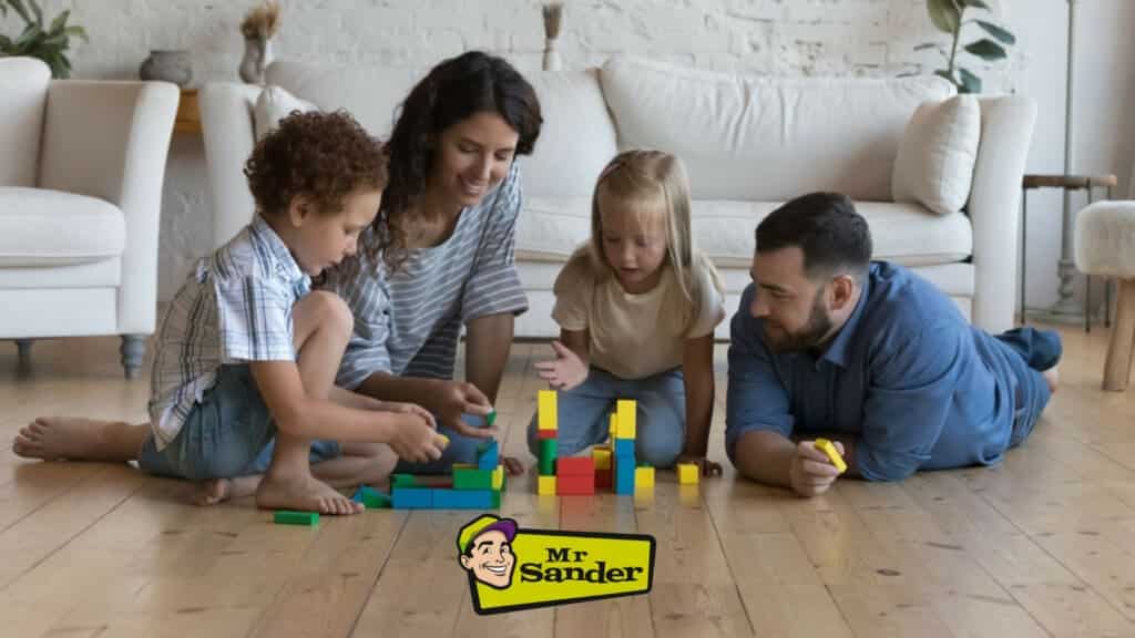 A happy family playing with colorful building blocks on the polished wooden floor of their living room in Hanwell, UK. The room features a comfortable white sofa and modern decor, highlighting the superior quality of Mr Sander® services in creating a beautiful and inviting living space with expertly restored and gleaming wooden floors.