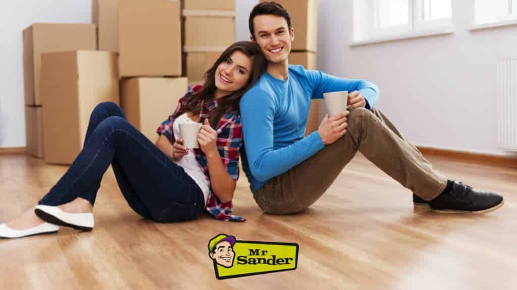 A happy couple sitting on their newly sanded and refinished oak wood flooring done by Mr Sander® in St Johns Wood, UK. They are leaning against each other with mugs in hand, surrounded by moving boxes. The Mr Sander® logo is visible, showcasing the professional floor restoration services provided. The bright room features large windows and a radiator, highlighting the beautifully restored oak flooring.