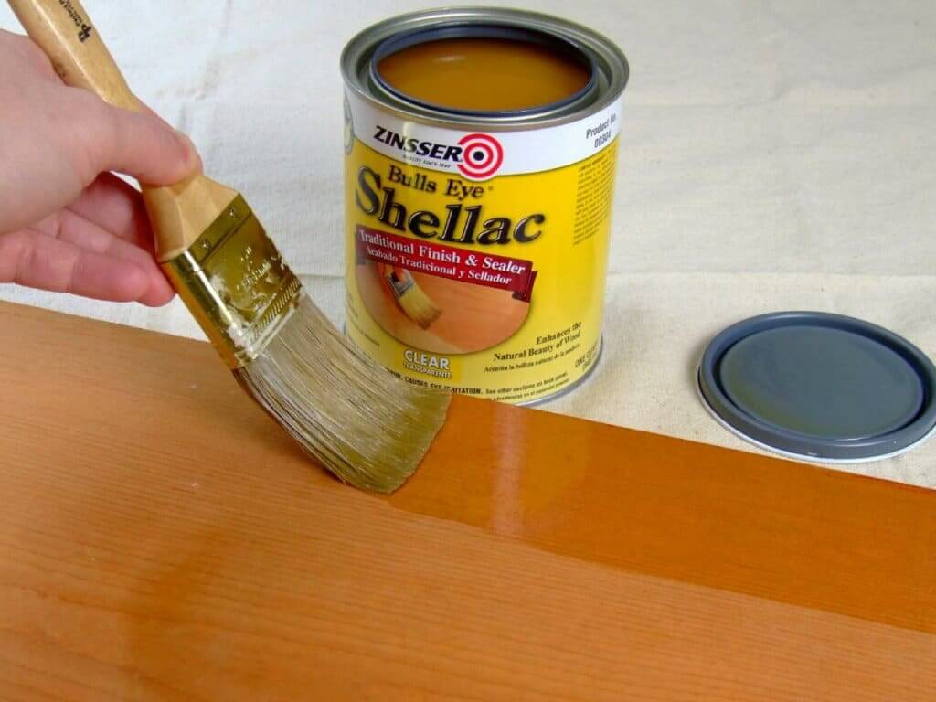 A person applying Zinsser Bulls Eye Shellac, a traditional finish and sealer, with a brush on a wooden surface. The can of shellac and its lid are placed nearby on the work surface.