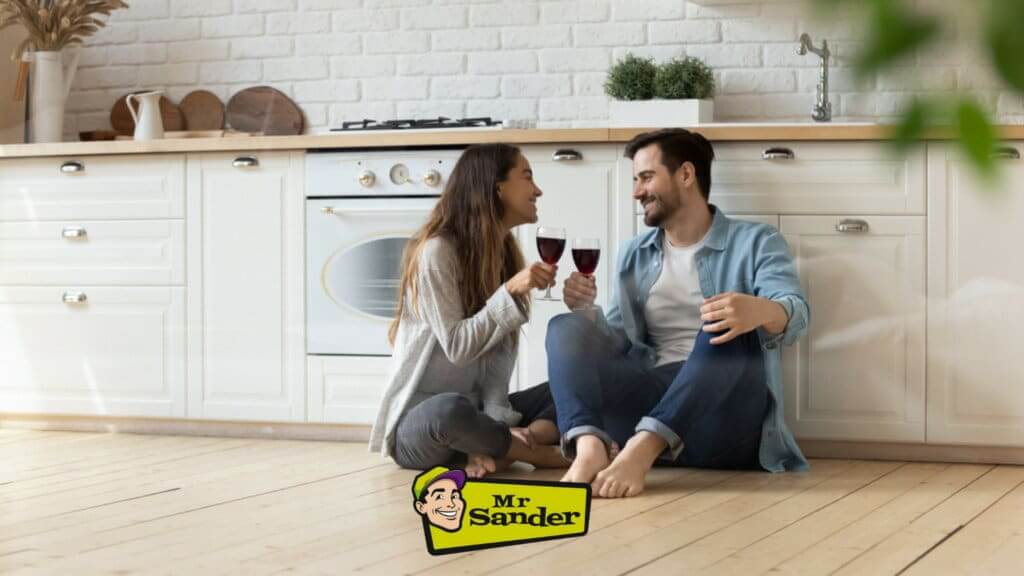 A happy couple sitting on a beautifully sanded and refinished wooden kitchen floor, done by Mr Sander®. They are smiling and toasting with glasses of red wine, enjoying the cozy ambiance of their white, modern kitchen with brick walls and potted plants.