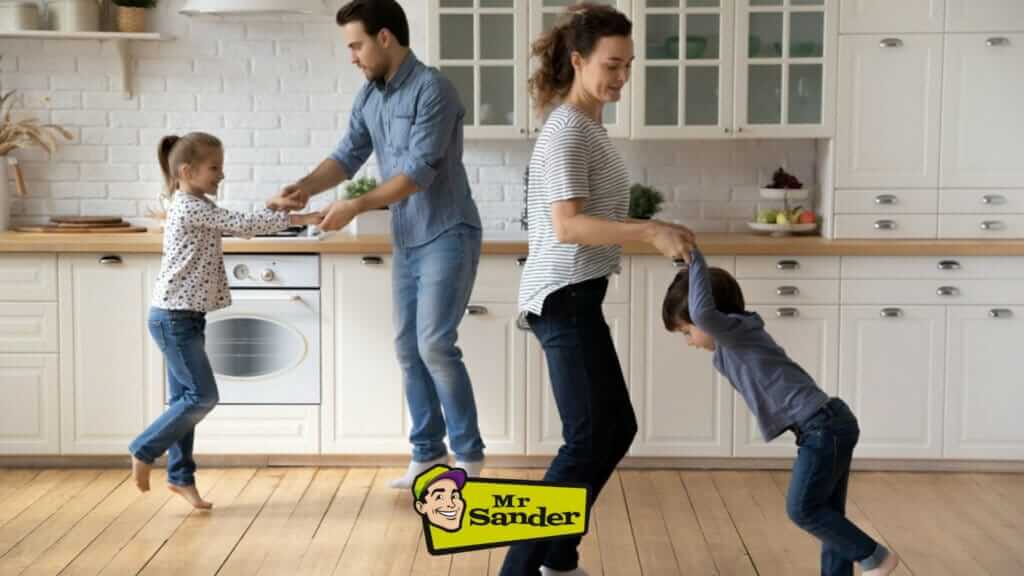 A joyful family of four dances in their kitchen on a beautifully sanded and refinished oak hardwood floor, showcasing the excellent work done by Mr Sander® in Burgess Hill, UK. The floor gleams under their feet, highlighting the smooth, flawless finish achieved through professional sanding and refinishing services.