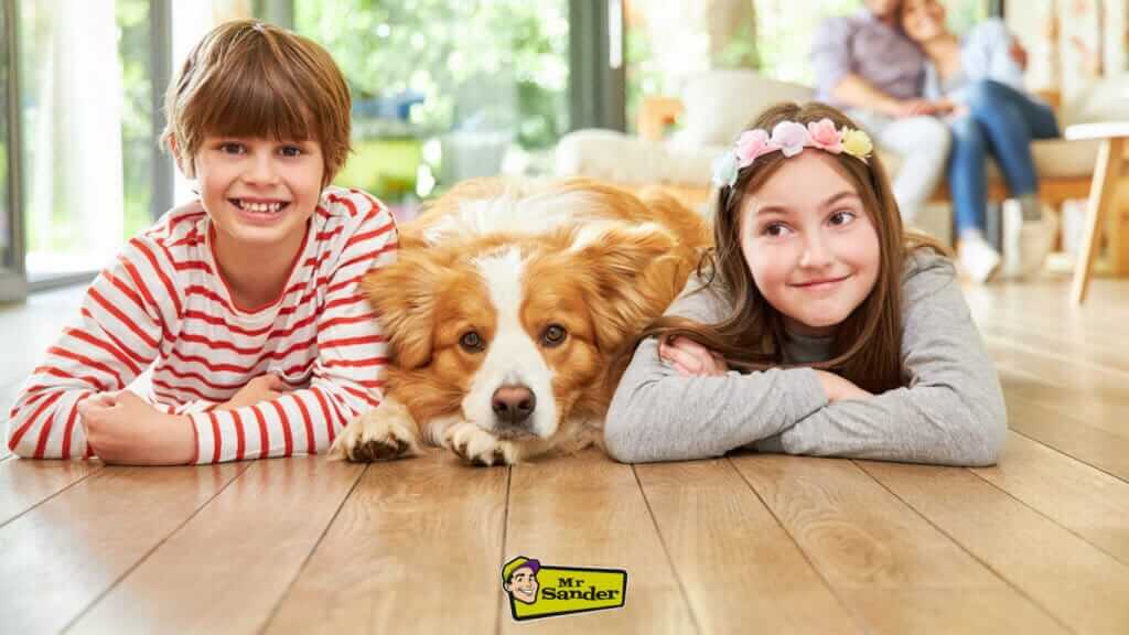 Two smiling children and a dog lying on a beautifully sanded and refinished wooden floor by Mr Sander®.