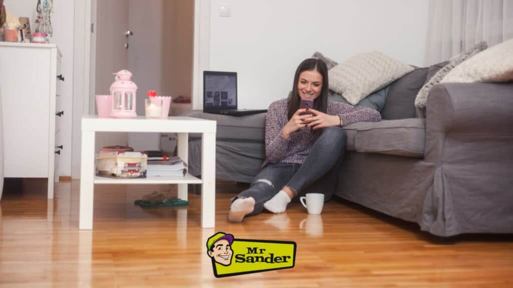 A woman sits comfortably on the floor next to a gray sofa, enjoying her phone in a cozy living room in Belvedere, UK. The room features a beautifully refinished oak hardwood floor, expertly sanded and polished by Mr Sander®, enhancing the warm and inviting atmosphere.
