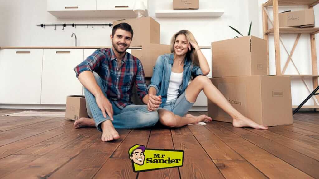 A smiling couple sitting on their newly refinished dark oak plank floor in their modern kitchen, surrounded by moving boxes. The floor sanding and refinishing were expertly done by Mr Sander® in Bermondsey, UK, transforming the space with rich, warm wood tones.