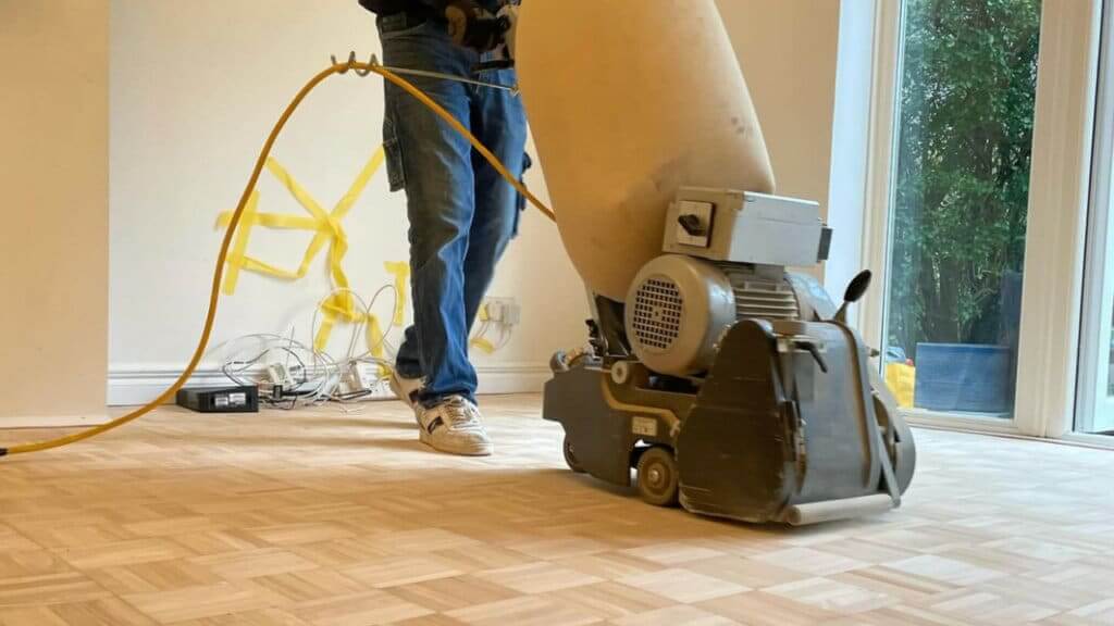 A professional from Mr Sander® is sanding a parquet floor in Professional Floor Sanding by Mr Sander® Using Frank Cobra Belt Sander in a Surrey Docks Living Room, UK, UK, using a Frank Cobra Belt Sander. The high-powered, dust-free machine weighs 78kg and features a 2500W motor, 2100rpm sanding speed, and 200x750 endless belt. The Frank Cobra offers quick belt changes, integrated DCS for 99.99% dust extraction, featherlite clutch control, and automatic belt adjustment. Ideal for professional sanding and stripping of all wooden floors, including parquet.