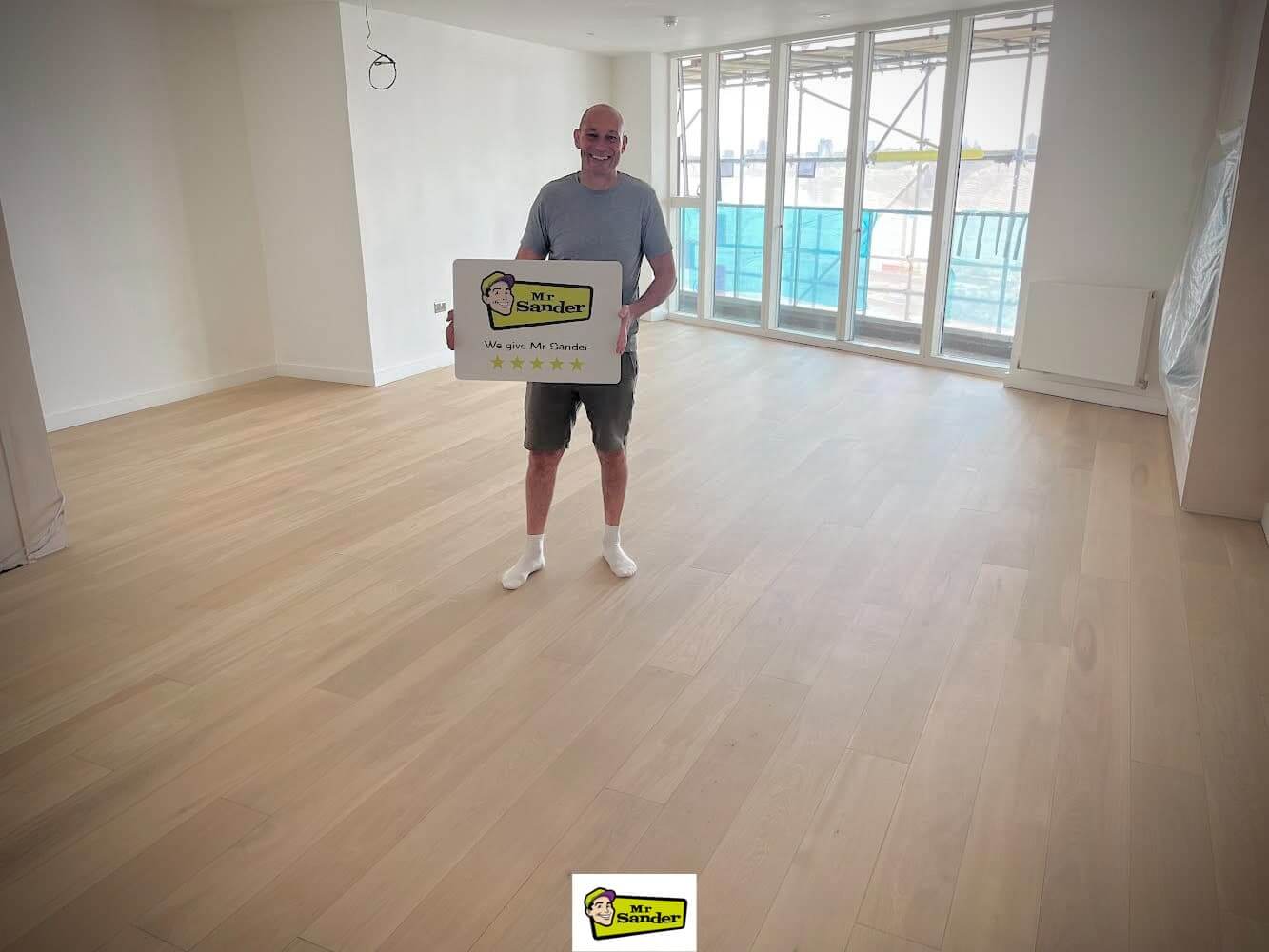 Man standing in a newly refinished living room with light wooden floors, holding a sign with Mr Sander® logo