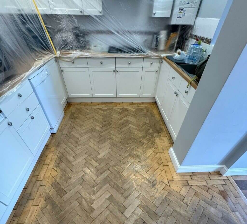 A kitchen with white cabinets and appliances, with plastic sheets protecting the countertops during a parquet flooring restoration. The floor shows visible wear and dirt, indicating the need for professional restoration. This image highlights the preparation and initial stages of the parquet flooring restoration process by Mr Sander®.