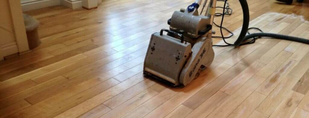A professional from Mr Sander® is sanding a solid oak hardwood in a living room in Burgess Hill, UK using a Bona Belt sander (AMO110000.3). The machine features a 2.2 kW motor, 230 V voltage, 50 Hz frequency, and a sanding belt size of 200 x 750 mm or 250 x 750 mm. The total weight is 74 kg.