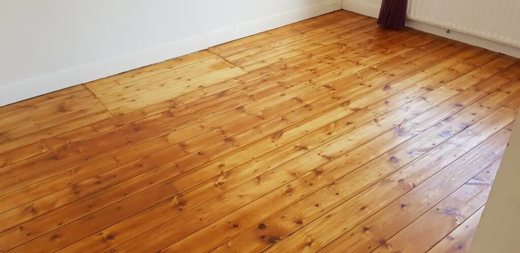 A room featuring a freshly sanded and refinished pine wood floor with a glossy finish. The floor showcases natural grain patterns, enhancing its warm, golden appearance. White skirting boards and a radiator are visible in the background.