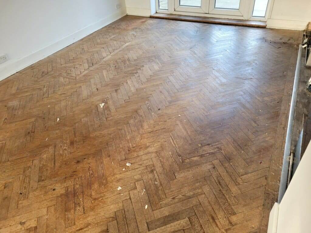 A living room with a worn and scuffed herringbone parquet floor before sanding and refinishing by Mr Sander®. The floor shows signs of heavy use, with visible scratches, stains, and uneven surfaces. The room has white walls and large glass doors leading outside.