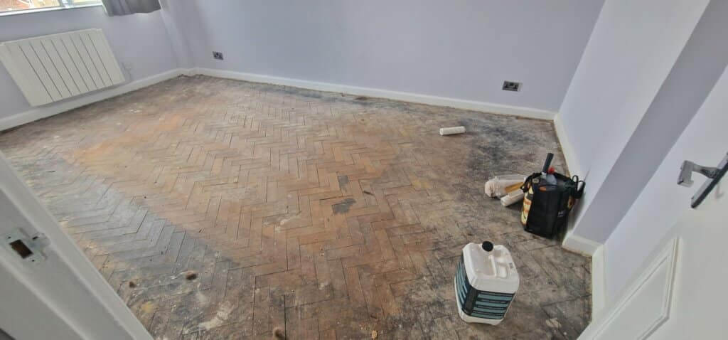 A room with an old, dusty parquet floor showing significant wear and tear, awaiting professional restoration. Various tools and materials are scattered around, indicating the preparation phase for parquet flooring restoration by Mr Sander®. This image highlights the initial state before the transformative restoration process begins.