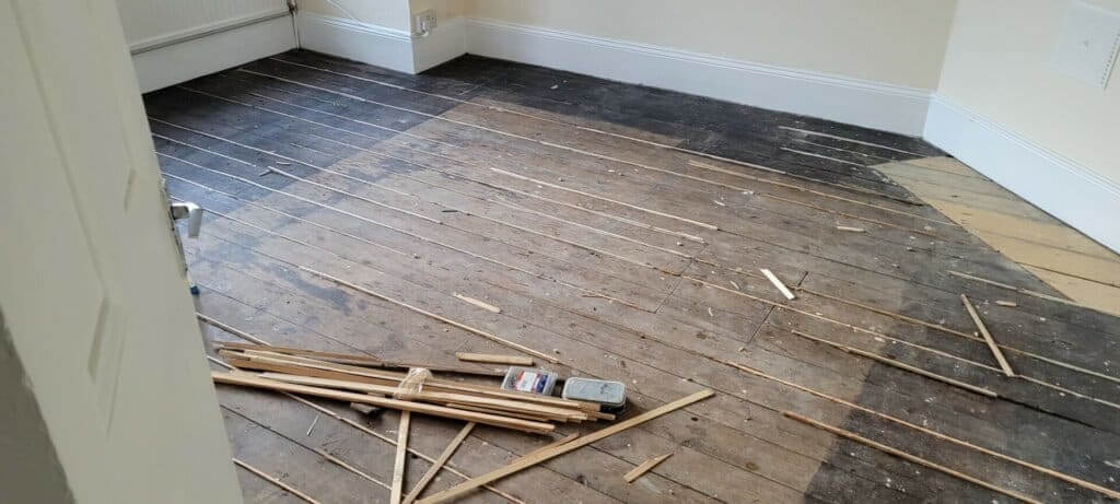 A room with a partially sanded wooden floor, showing areas of dark, unfinished wood and lighter, sanded sections. Wooden strips and tools are scattered on the floor, indicating ongoing floor sanding and refinishing work.