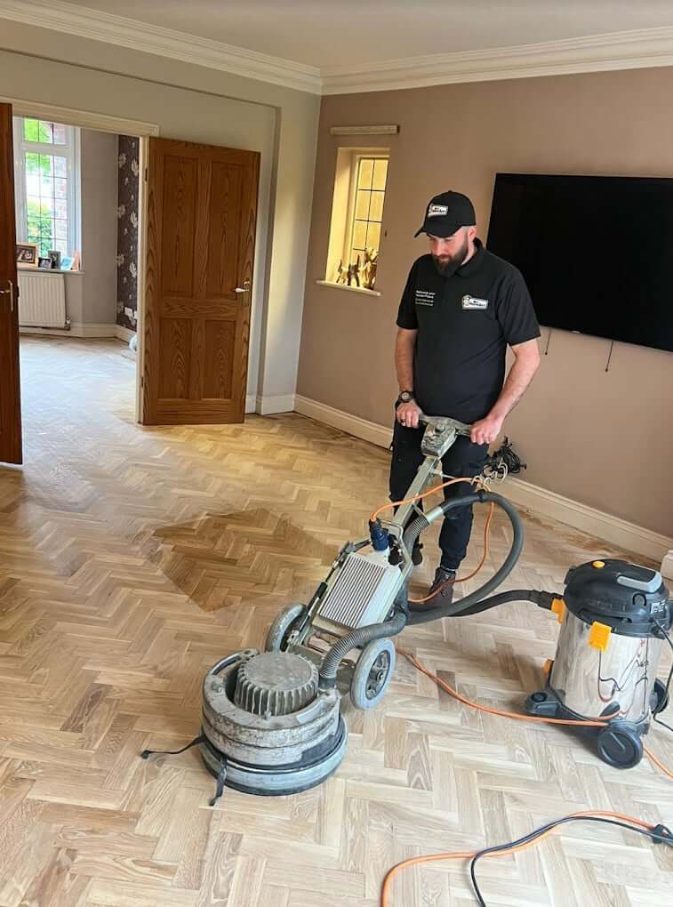 A man from Mr Sander® sanding a parquet floor in a living room in Old Street, UK, using the PALLMANN SPIDER wood floor sanding machine. The PALLMANN SPIDER features 360° illumination, switchable power reserve, triple-disc technology, infinite speed adjustment (50-400 RPM), a 230 V power supply, 2000 W power consumption, 400 mm sanding width, 42 kg weight, and an additional weight of 15 kg for increased sanding pressure.