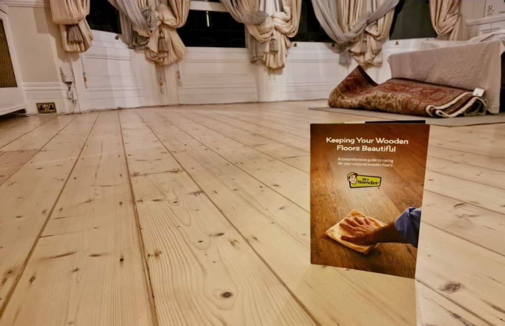 A beautifully refinished light pine wood floor by Mr Sander® in UK. The floor features smooth planks with a natural finish, and in the foreground, there is a brochure titled "Keeping Your Wooden Floors Beautiful," offering care tips for the restored wooden floors. The room has elegant drapery and a cozy atmosphere with rugs placed in the background.