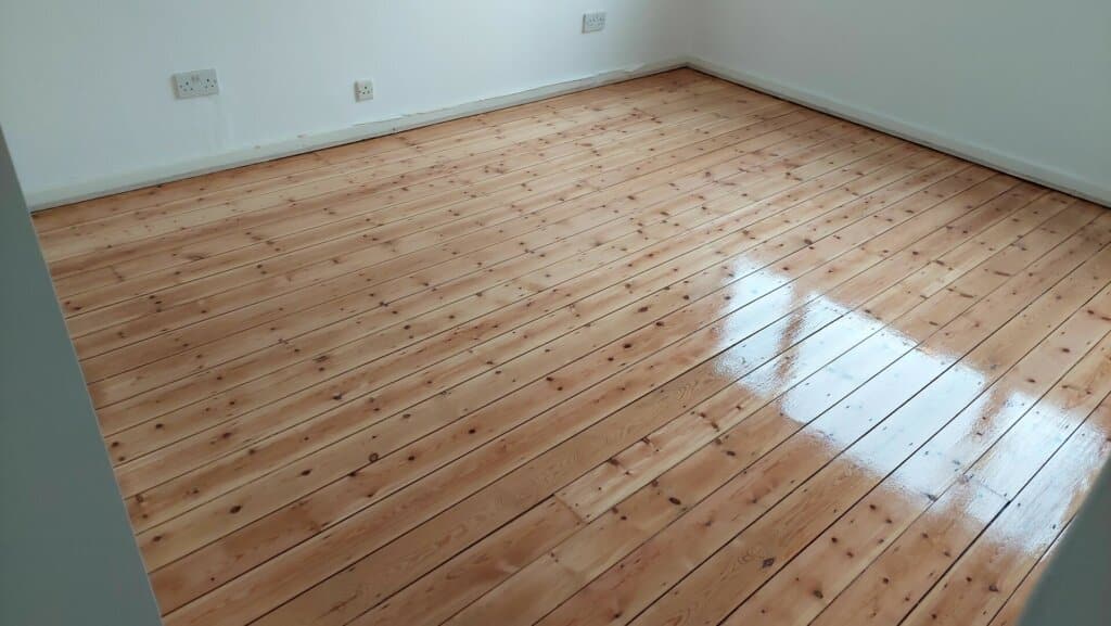 A freshly sanded and refinished pine wood floor in a bright, empty room with white walls. The floorboards are polished to a high shine, reflecting light from a nearby window.
