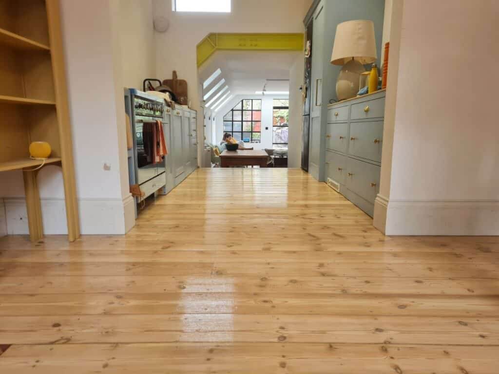 A beautifully sanded and varnished pine wood floor in a cozy, modern home. The floor extends through an open-plan kitchen and dining area, with light-colored cabinetry and a window at the far end letting in natural light. The space is warm and inviting, showcasing the expert craftsmanship of Mr. Sander.