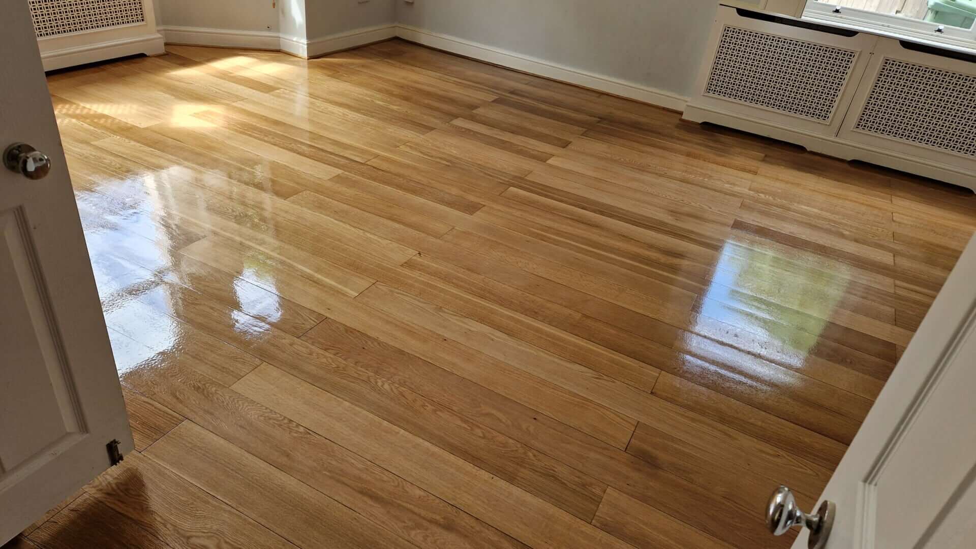 A bright room with a newly sanded and refinished wooden floor that has a high-gloss finish. The sunlight from the window reflects off the polished surface, highlighting the smooth and even application of the finish.