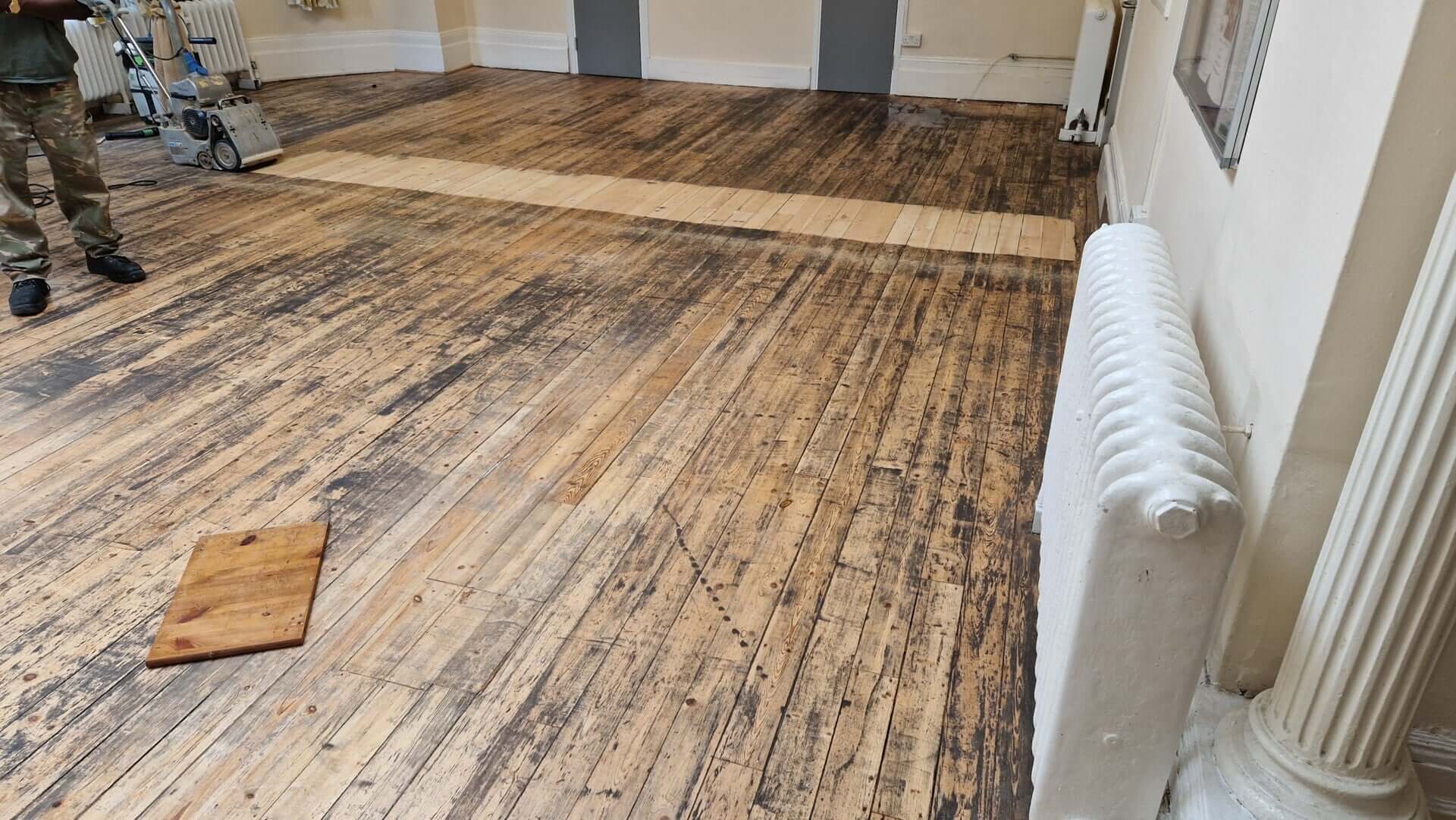 A room with a partially sanded and refinished pine wood floor, showing a stark contrast between the newly sanded section and the old, worn section. A sanding machine is visible, along with a worker in camouflage pants.