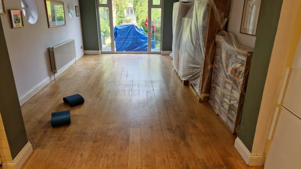 A living room with a partially sanded oak wood floor. Furniture covered with plastic sheets is lined up against the wall, and there are yoga mats on the floor, indicating ongoing floor refinishing work. The room has large windows leading to a garden.