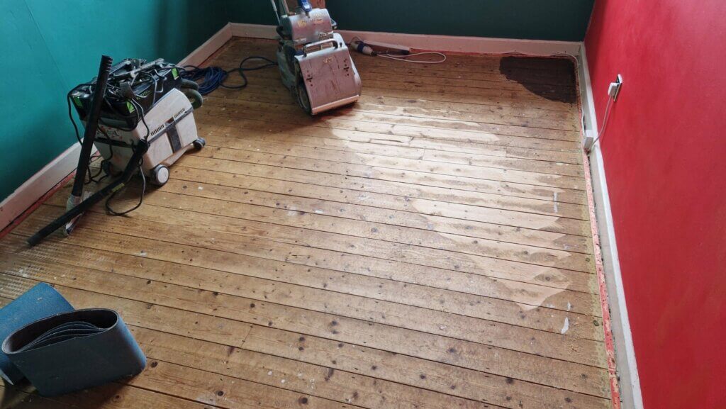 A living room in Hatfield, UK, where a Mr Sander® professional is sanding an oak wood floor using a Bona Belt (AMO110000.3) belt sander. The machine features a 2.2 kW motor, 230 V voltage, 50 Hz frequency, with a drum width of 200 and 250 mm, and a sanding belt size of 200 x 750 mm or 250 x 750 mm. The sander's total weight is 74 kg. The room has green and red walls, showcasing the floor restoration in progress.