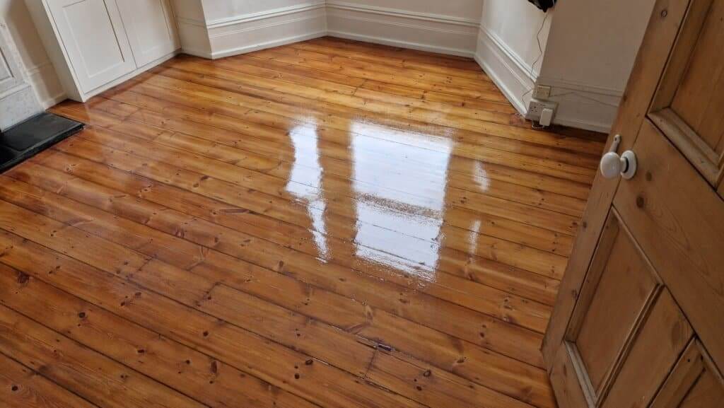 A freshly restored wooden floor with a glossy finish, reflecting light from a nearby window. The floorboards have a warm, polished appearance, enhancing the room's natural charm.