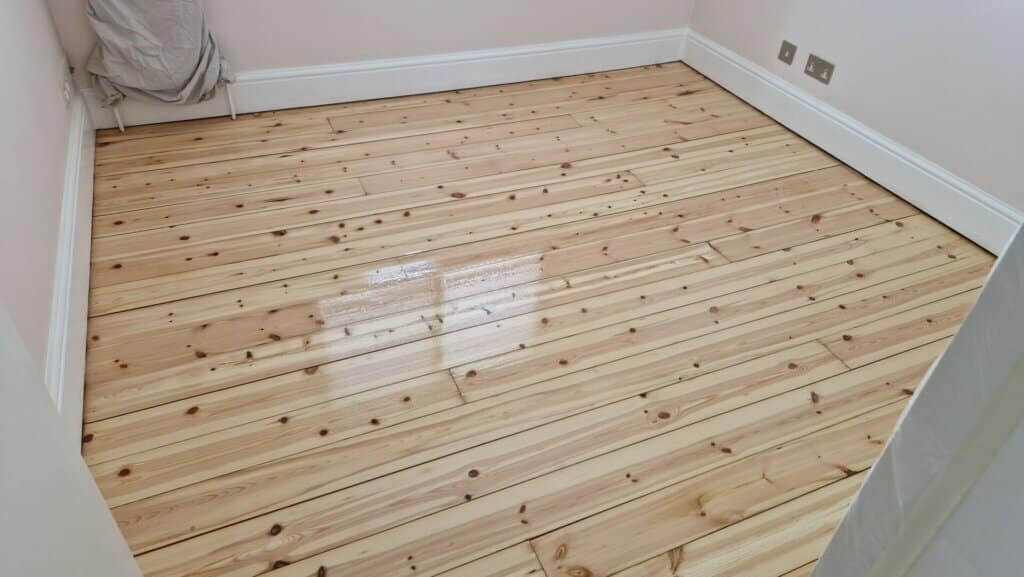 A newly refinished wooden floor with a glossy finish in a small, well-lit room. The natural wood grain and knots are visible, giving the room a warm and inviting look.