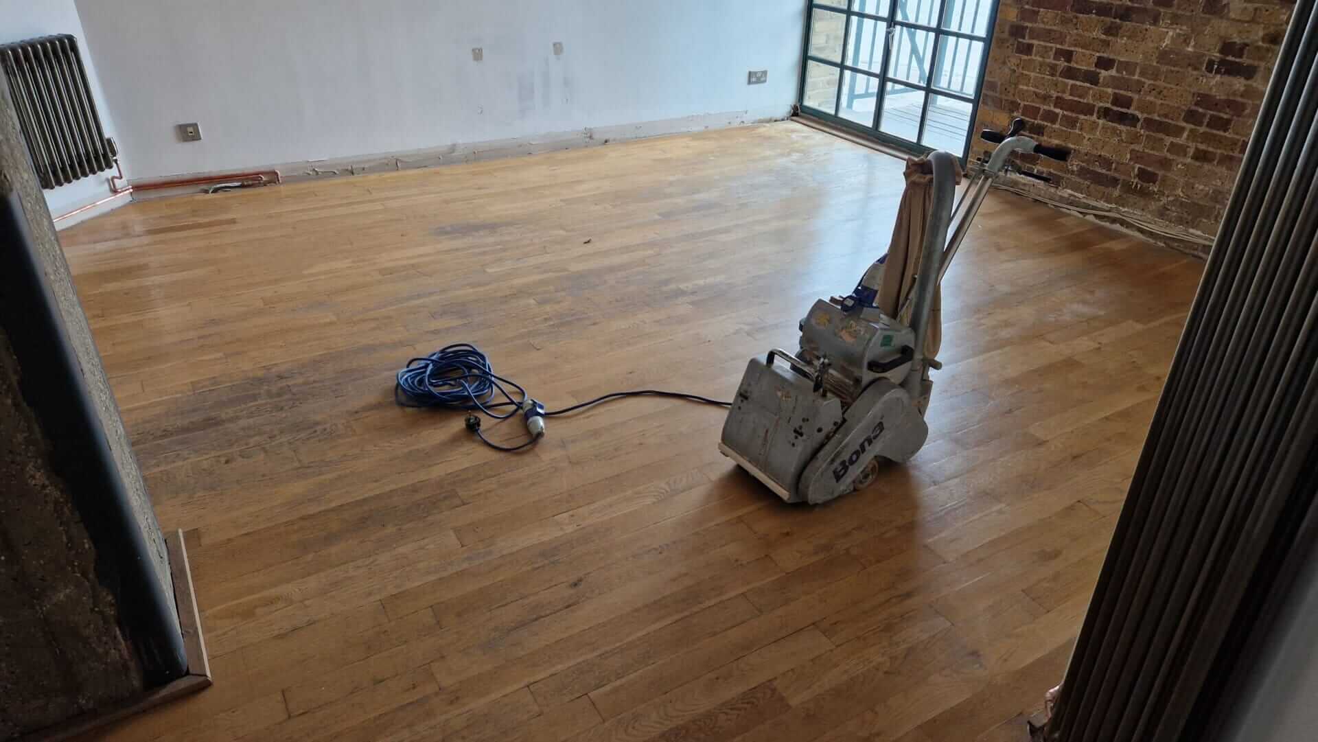 Mr. Sander® technician using a Bona Belt sander (AMO110000.3) with 2.2 kW motor power, 230 V voltage, and 50 Hz frequency, sanding a floor in a modern UK living room. The sander features a drum width of 200 & 250 mm, sanding belt size of 200 x 750 mm or 250 x 750 mm, and weighs 74 kg.