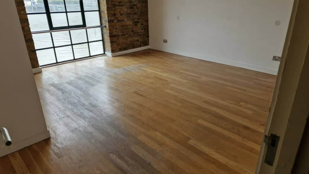 A spacious room with large windows and a brick accent wall. The floor is partially refinished, with a clear distinction between the sanded and unsanded sections. The natural wood grain of the floorboards is visible, and the windows provide a view of an outdoor water scene. The room is empty, highlighting the floor's condition.