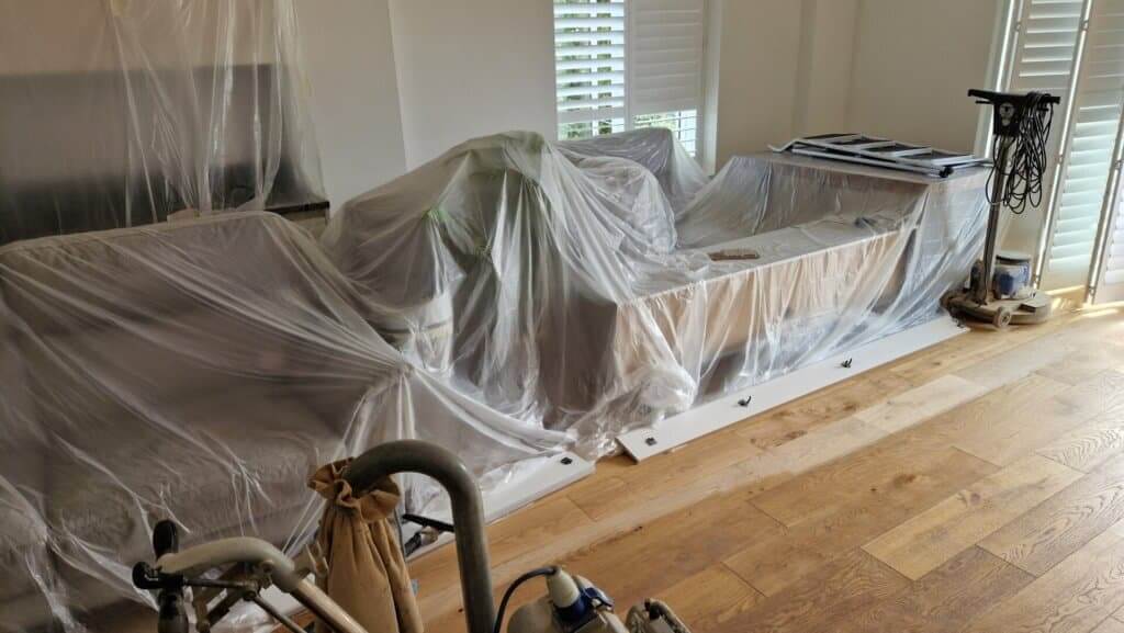 A living room prepared for floor sanding and refinishing, showcasing protective plastic sheeting over the furniture and appliances. The area features unfinished oak hardwood floors, highlighting the meticulous care taken to safeguard the environment during the sanding process. Equipment is visible in the foreground, emphasizing the professional approach by Mr. Sander® in maintaining the beauty of the hardwood flooring.