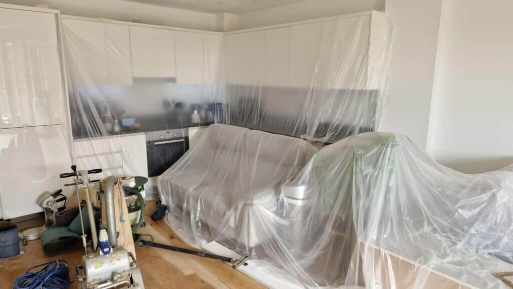 A kitchen area undergoing preparation for floor sanding and refinishing by Mr. Sander®. The space is covered with plastic sheeting to protect the cabinets and furniture while sanding equipment is visible in the foreground. The polished engineered hardwood floors are partially exposed, showcasing the meticulous care taken to protect the surrounding environment during the refinishing process.