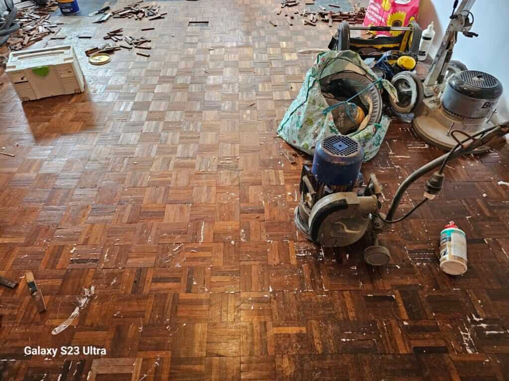 A partially sanded wooden parquet floor undergoing restoration. Various tools, including floor sanders, vacuum hoses, and wood pieces, are scattered across the room. The floor shows signs of wear and old adhesive, indicating the beginning stages of the sanding and refinishing process.