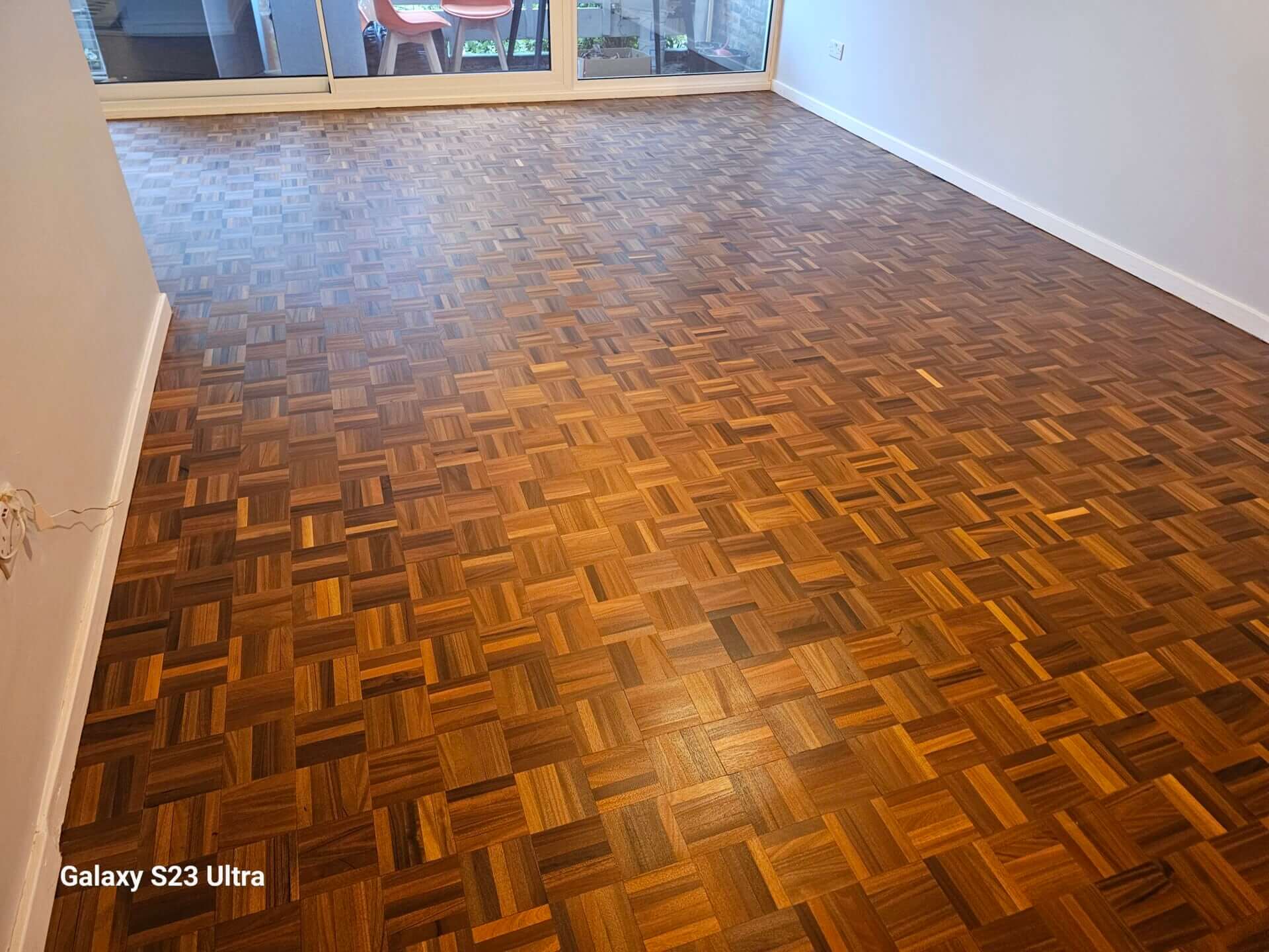 A beautifully refinished parquet wood floor in a spacious room with large glass doors leading to a balcony. The rich wood tones of the parquet squares are enhanced by the polished, smooth finish, giving the room a warm and inviting feel. The floor sanding and refinishing were expertly done by Mr Sander®.