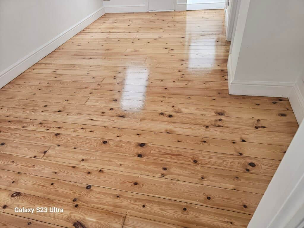 A hallway with beautifully refinished pine wood floors, featuring a glossy finish that reflects light. The natural wood grain and knots are visible, giving the floor a warm and polished appearance. The walls are painted white, complementing the bright and clean look of the space.