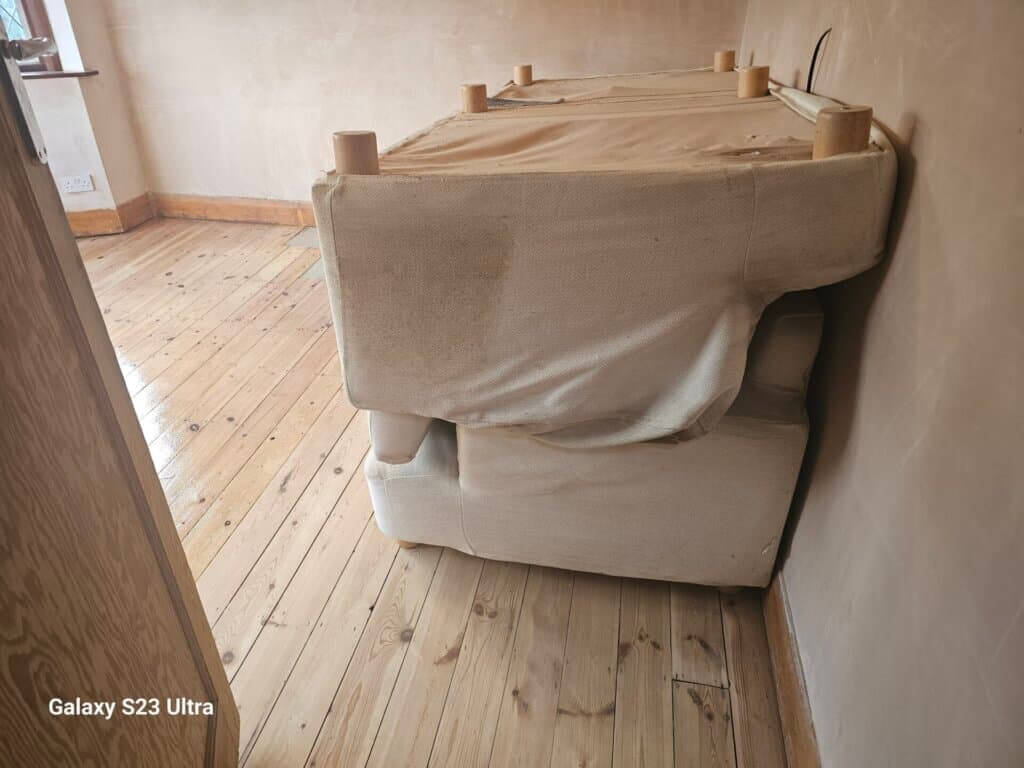 A room with freshly sanded and refinished light pine wood floors, featuring natural knots and a glossy finish. A couch, wrapped in protective fabric, is stacked to the side of the room. The walls are unpainted, indicating ongoing renovation work.