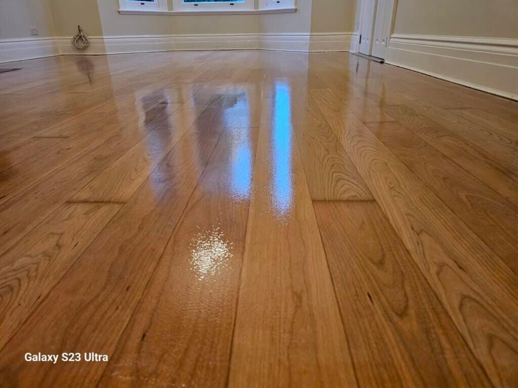 A close-up of a beautifully refinished hardwood floor with a glossy finish, reflecting light from nearby windows. The floor features a smooth, polished surface with visible wood grain patterns. The baseboards and walls are painted a light color, highlighting the elegance of the restored floor.