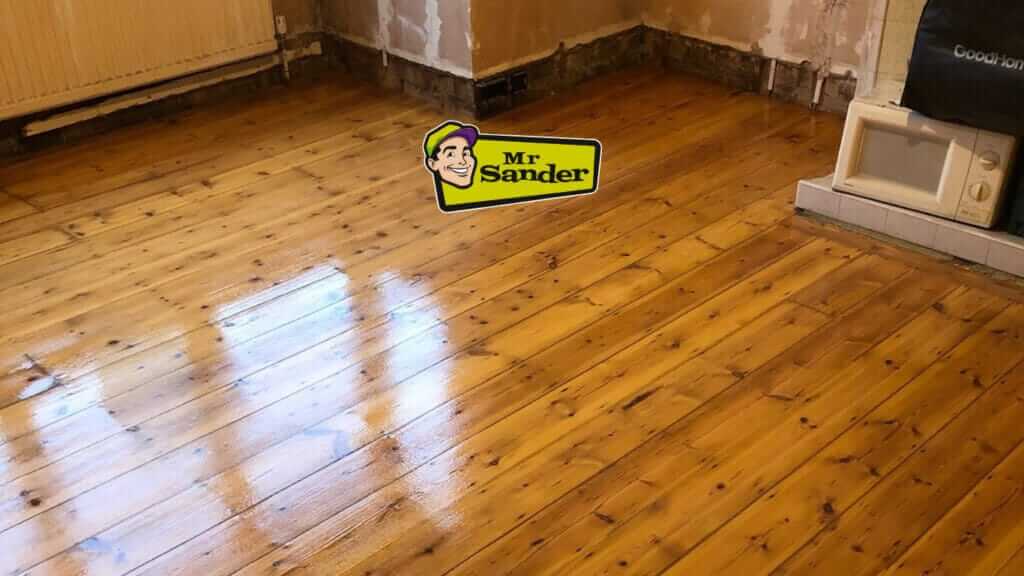A close-up view of a beautifully restored pine floor in a living room. The floorboards are polished and shiny, with sunlight streaming in and casting shadows across the surface.