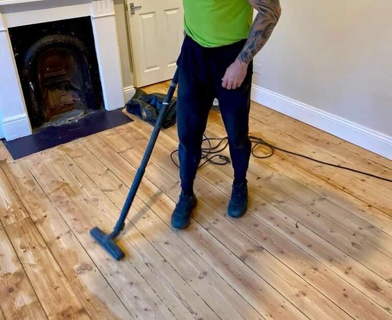 A worker in a green shirt using a vacuum to clean up sawdust from a newly sanded wooden floor, preparing it for refinishing.